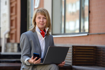 Portrait of a mature business woman with a smartphone and a laptop.  Works outdoors, near the office. 