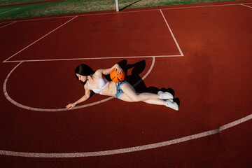 Young athletic female, in top and sweatpants, playing with ball on basketball court outdoors.