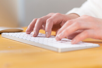 An image of a woman's hand is typing on a white keyboard on a wooden table. Selective focus