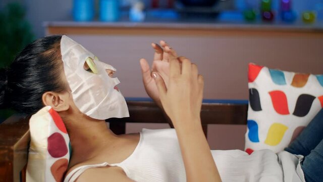 Relax and spa concept - Lady with face mask putting cucumber slices on her eyes. Closeup shot of a lady wearing a serum sheet mask while resting on the sofa - skincare at home concept