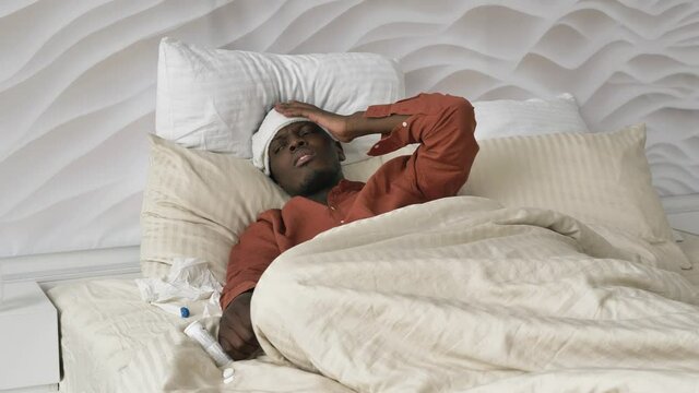 Upset African-American man with patch on head pills and thermometer suffers from cold lying on large bed at home close view