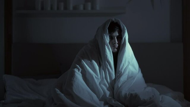 Night insomnia. Trouble sleeping. Anxiety fatigue. Loneliness depression. Tired exhausted sad sleepy man in blanket sitting in bed alone late in dark bedroom.