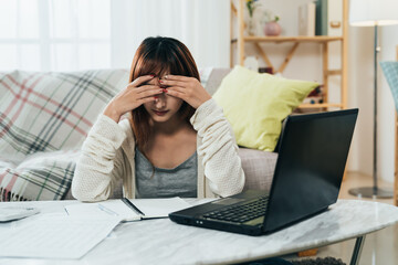 front view asian girl sitting and burying her head in hands is worried about debt while filing tax with a laptop in the living room at home.