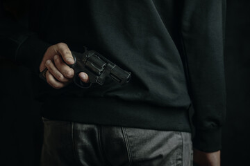 Man in grey cloth holds firearm behind his back. Revolver in men's hand. Person ready to attack with hand gun. Criminal concept.