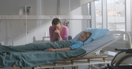 Sick little boy lying in hospital bed sleeping, his mother worries and prays beside him.
