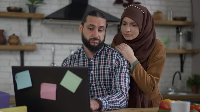 Portrait of serious successful Middle Eastern businessman typing on laptop keyboard in kitchen as irritated woman in hijab talking and gesturing. Overworking husband ignoring sad young beautiful wife
