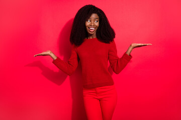 Photo of shiny cute dark skin lady wear casual sweater smiling compare arms empty space isolated red color background
