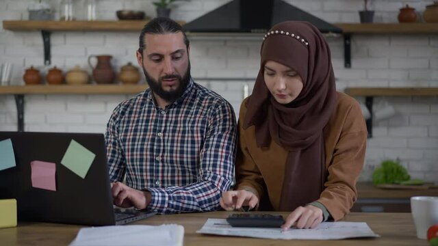 Concentrated Middle Eastern wife calculating family budget as focused husband working online on laptop in kitchen. Muslim couple on remote working in home office indoors