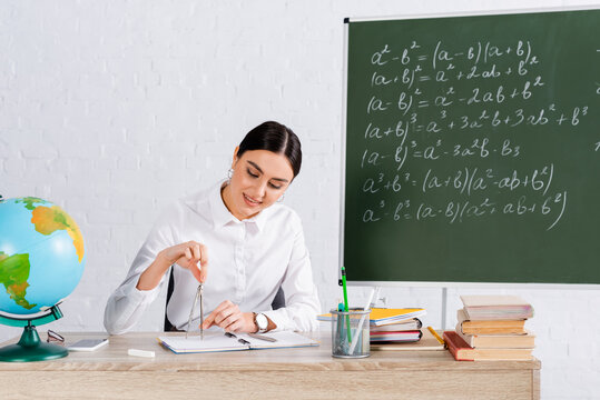 Smiling teacher holding drawing compass near books and notebooks on table - Powered by Adobe