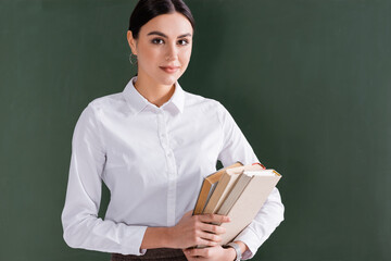 Brunette teacher with books looking at camera near chalkboard