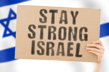 The phrase " Stay strong Israel " on a banner in men's hand with blurred Israeli flag on the background. Support. Patriot. Community. Free. Freedom. Independence. Nation. Problem. Issue