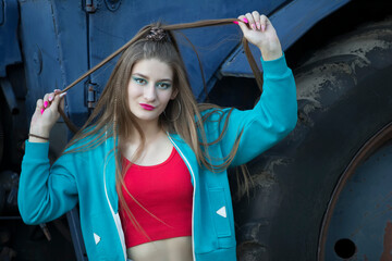 A beautiful country girl in the style of the 90s in bright clothes stands near a tractor.