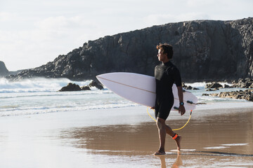 Young surfer walking by the beach shore watching the waves holding surfboard