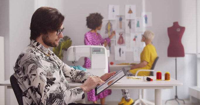 Young male fashion designer use tablet in office with colleagues discussing on background - Powered by Adobe