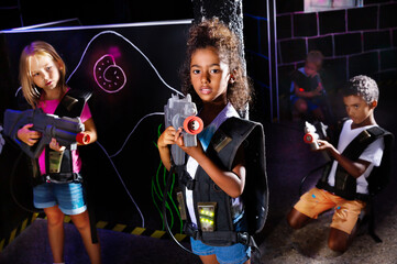 Fototapeta na wymiar Portrait of african preteen girl standing with a laser gun on dark lasertag arena on background with her team