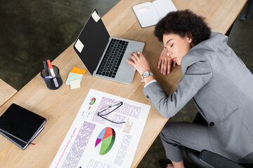 High angle view of african american businesswoman lying with closed eyes near laptop and paper on table