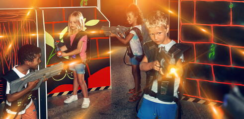 Modern preteen girls and boys of different nationalities with laser pistols playing laser tag on the dark labyrinth