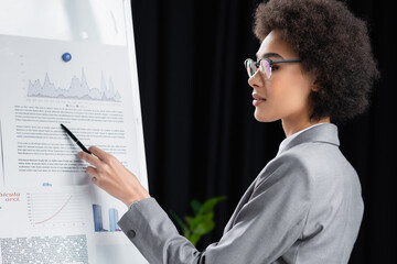 Side view of young african american businesswoman with pen pointing at paper on flipchart