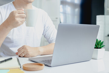 Man in a white shirt picks up a coffee mug and thinks of work on his laptop while working at home.
