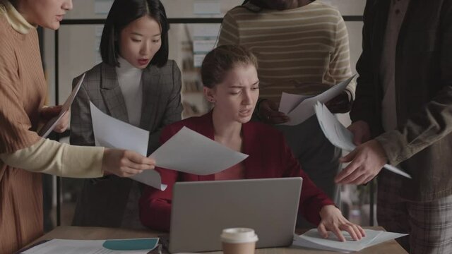 Chest-up of young Caucasian businesswoman sitting by desk by laptop computer, overwhelmed by employees loading her with paperwork