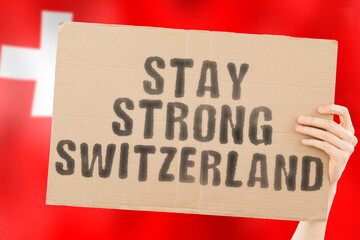 The phrase " Stay strong Switzerland " on a banner in men's hand with blurred Swiss flag on the background. Support. Patriot. Community. Free. Freedom. Independence. Nation. Problem. Issue