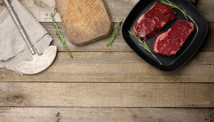 two raw pieces of beef in a black square grill pan, cutting board and knife on a wooden table