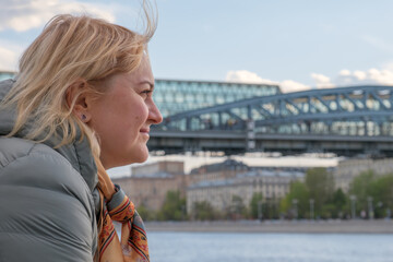 Blonde middle-aged woman in profile stands on embankment and looks at river against the background of city bridge. Wind develops the woman's hair. City walks, reasoning about life.