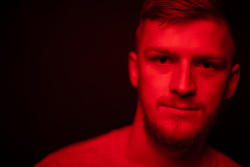 Young attractive male person posing in contrast red light. Portrait of handsome caucasian guy in dark studio with red neon lightning. Confident man looks at camera, fashion beauty concept