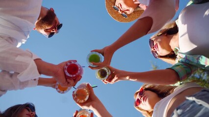 Low angle view of friends having fun at pool party, clinking glasses with colorful summer cocktails near hotel swimming pool. People toast drinking fresh juice at luxury summer villa in slow motion. - Powered by Adobe