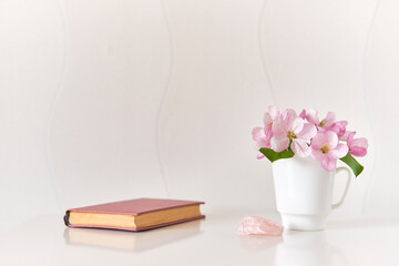 Gentle bouquet of pink apple tree flowers in a white coffee cup, pink mineral, small poetry book on a white coffee table