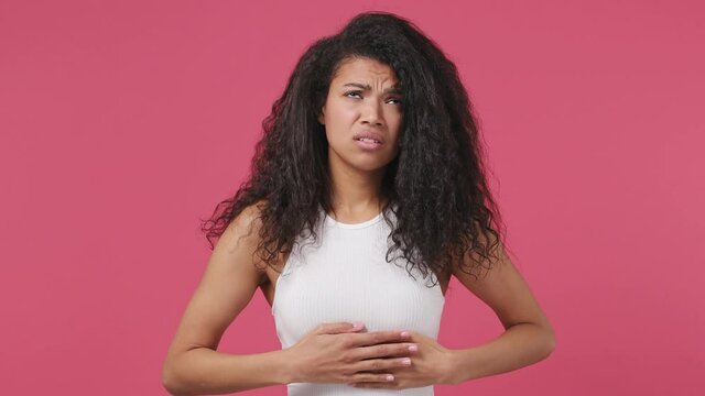 Sick ill sad tired young african woman wears white tank top shirt put hands on abdomen suffering from stomach-ache griping bellyache feel bad seedy isolated on dark pink color wall background studio