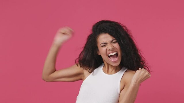 Excited overjoyed happy young african woman curly hair 20s years old wear white tank top shirt doing winner gesture celebrate clenching fists say yes isolated on dark pink color wall background studio