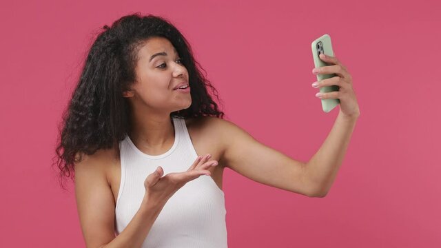 Young fun african woman wears white tank top shirt get video call using mobile cell phone doing selfie talk conducting pleasant conversation greet with hand isolated on dark pink color wall background
