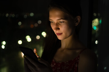 Portrait of a beautiful young woman in a red dress and with red lipstick on her lips standing near the window at night holding a mobile phone near her face, soft light from the phone falls on her face