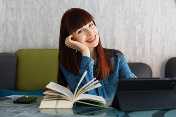 Smiling happy pretty young girl in jeans dress, enjoying her work and leisure time, sitting at the table with open book and tablet pc. People, emotions, gadgets and lifestyle concept