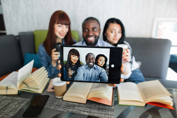 Young cheerful multiracial friends taking selfie in cafe or college campus, using digital tablet. Multiethnic students, sitting at the table with books and making self portrait on tablet pc