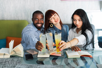 Jolly three friends clinking glasses of tasty cocktails in cafe. African man, Caucasian and Asian women, enjoying study time together, reading books and drinking juice in modern coffee shop