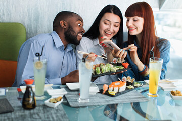 People, gadgets, food and blogging concept. Team of multicultural young people in cafe eating sushi rolls, having fun. Red haired Caucasian woman makes a photo of sushi on phone