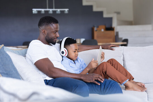 African american father and son sitting on sofa, using tablet and smiling