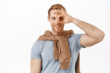 Happy attractive redhead man showing okay OK sign over eye, smiling and looking pleased, praise something good, recommending great thing, say alright like gesture, white background