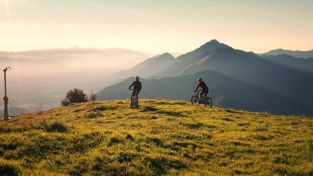 Aerial shot of two female mountain bikers riding across the meadow, stopping at the top of the hill, giving high five
