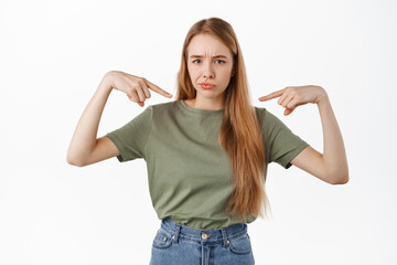 Sad and disappointed young woman pointing at herself, sulking from unfair situation, standing...