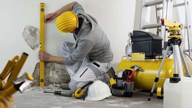 House Renovation Concept, Man Worker Looks At The Spirit Level Checks The Wall, Helmet And Safety Headphones, Air Compressor And Construction Tools In Background