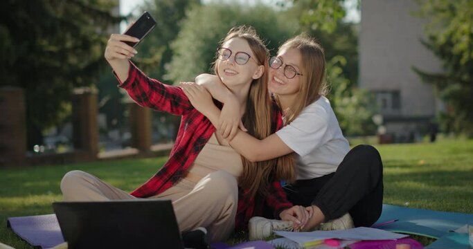 Taking a photo taking a selfie on the phone Two happy girls best friends sitting on the ground green grass hugging fun smiling
