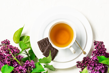 Artistic composition with cup of herbal tea, chocolate pieces and aromatic lilac flowers on white background top view. 