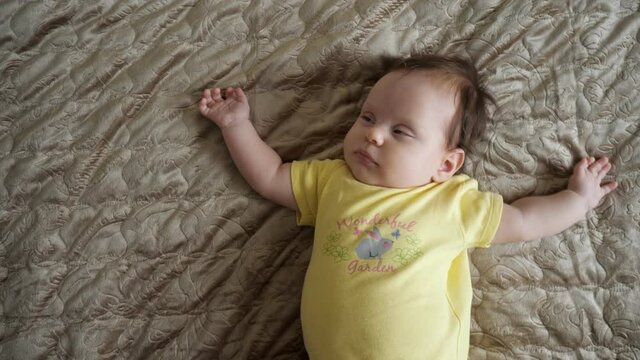 Baby girl wearing yellow clothing with picture awakes on large parents bed blanket grimaces and shape hands in apartment room close upper view