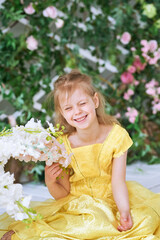Girl, 7, sits against a green wall with flowers in a yellow dress and sniffs a white flower