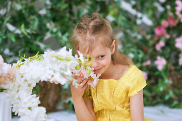 Fototapeta premium Girl, 7, sits against a green wall with flowers in a yellow dress and sniffs a white flower
