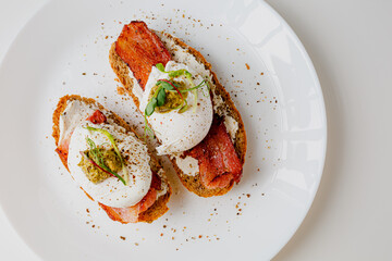 Avocado eggs benedict with pesto sauce, grilled bacon, pepper, cream cheese Philadelphia. Poached eggs on healthy bread toast with hollandaise sauce. Cut egg. The yolk flows out. Micro greens. 