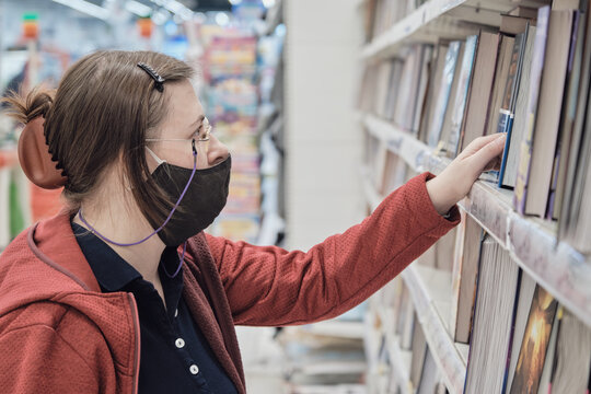 Serious caucasian woman wearing glasses with a chain and a mask against the virus. She buys a book in the second hand bookstore. Concept of reading literature and gaining new knowledge